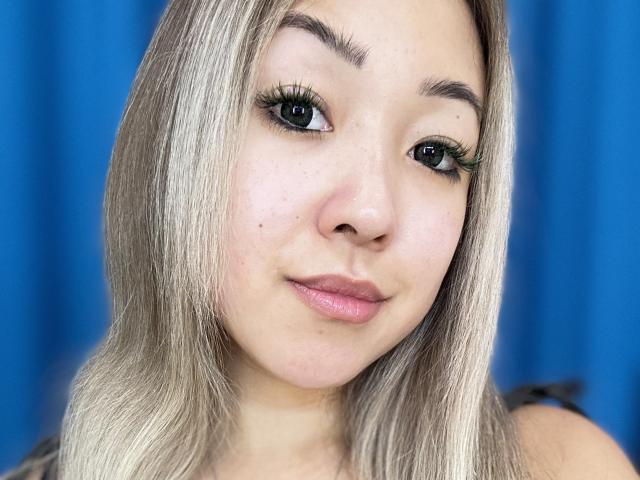 RenyLime has blonde, straight hair, green eyeliner, and long eyelashes. She's looking at the camera with a slight, pink-lipsticked smile. Her skin is fair with small moles.