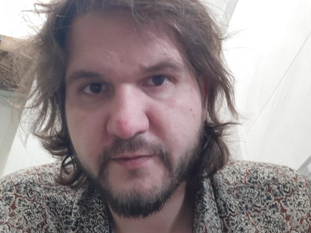 NectarineFad has a chubby face, brown wavy hair, a beard, and a slightly scruffy look. He's wearing a patterned shirt with a mix of brown and black.