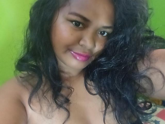 ChellaLove with curly black hair, pink glossy lips, and dark eyebrows, looking at the camera with a slight smile, showing her bare shoulders and upper chest.