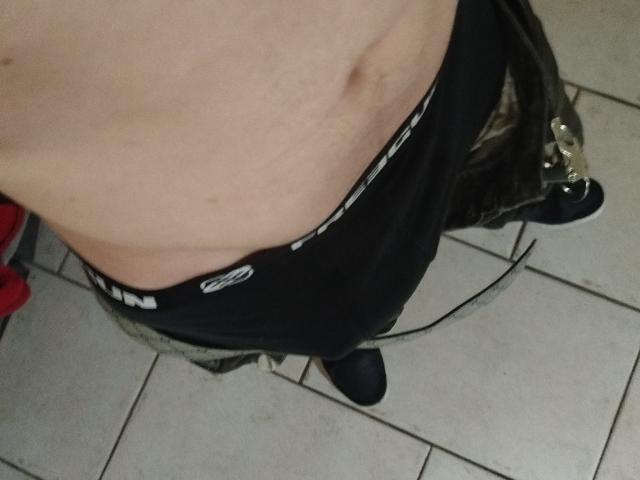 Gourmand69s' bare, slightly chubby belly with a small navel, wearing black underwear and unbuttoned jeans, standing on white tiled floor. Lower belly and black shoes visible.