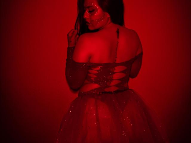 BellaSmithX69 in a red dress with cut-out back, sparkling fabric, and red lighting. Her ass is visible, and she's looking over her shoulder with dark hair.
