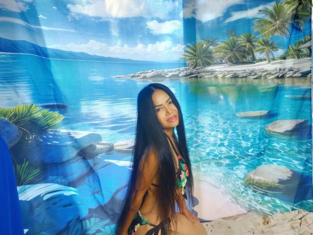 Lotusblume-hot with long black hair, wearing a colorful bikini, showing her ass, standing by a blue-tinted, tropical-themed mural with palm trees and clear water.