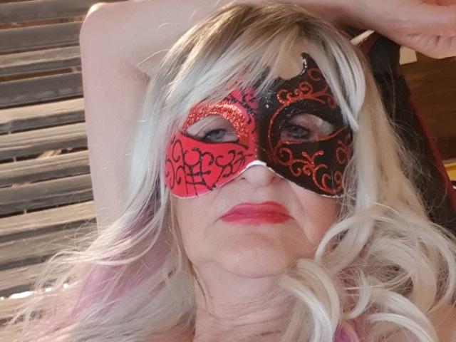 LadyHazel, con cabello rubio y ondulado, lleva una máscara de carnaval roja y negra con diseños elaborados, está recostada con los brazos por encima de la cabeza, tiene lápiz labial rojo y una expresión ligeramente seria..