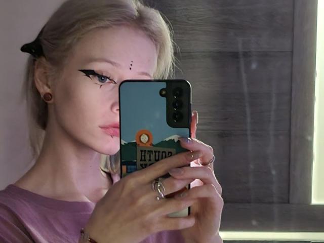 ExcitedBlizzard with blonde hair, black eyeliner, and small nose stud, holding a phone with a mountain and sun sticker, wearing a pink shirt, and multiple rings.