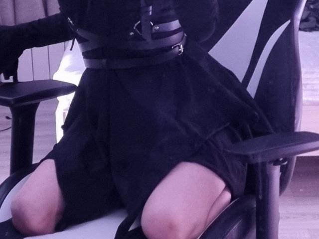 Der schwarze Rock von ExcitedBlizzard ist hochgezogen, wodurch blasse Oberschenkel sichtbar werden. Sie sitzt auf einem schwarz-weißen Stuhl mit schwarzen Trägern, die über ihre Taille verlaufen. Das Bild hat einen leicht violetten Farbton..