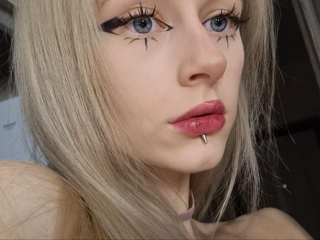 ExcitedBlizzard heeft blond haar, blauwe ogen, zwarte eyeliner met vleugels, roze lippenstift, een zilveren lipring en een choker. Haar gezicht is in het midden van de foto geplaatst en ze kijkt iets naar beneden..