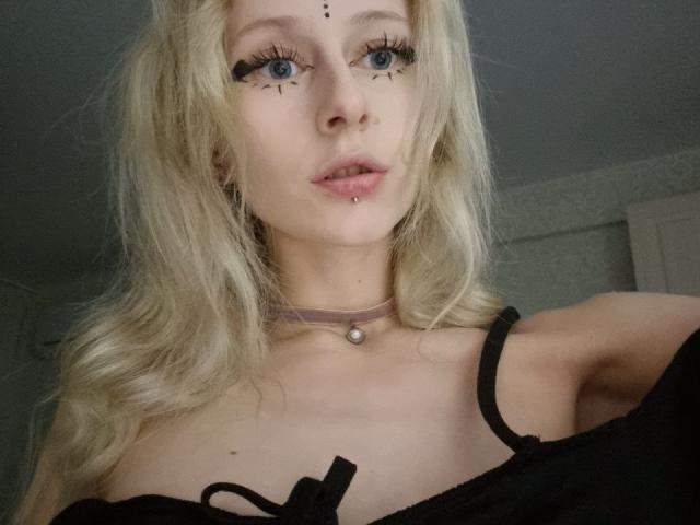 ExcitedBlizzard, com cabelo loiro, olhos azuis, delineador preto, piercing no nariz, piercing no lábio, colar, blusa preta com um ombro descoberto, mostrando os seios, em um selfie..