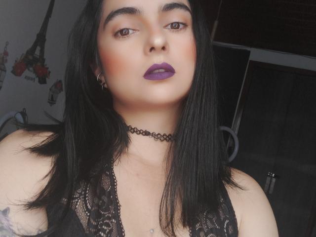 CandyParis with black lace top, purple lipstick, choker, and straight black hair, showing off her tits and tattoo, with a sexy, confident look.