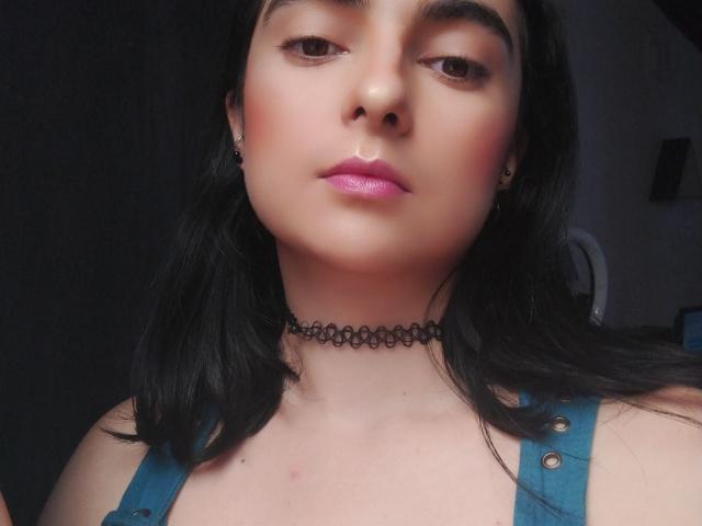 CandyParis heeft bleke huid, zwart haar, roze lippenstift, een zwarte choker en blauwe braces. Ze kijkt neutraal naar de camera. Haar borsten zijn licht zichtbaar..