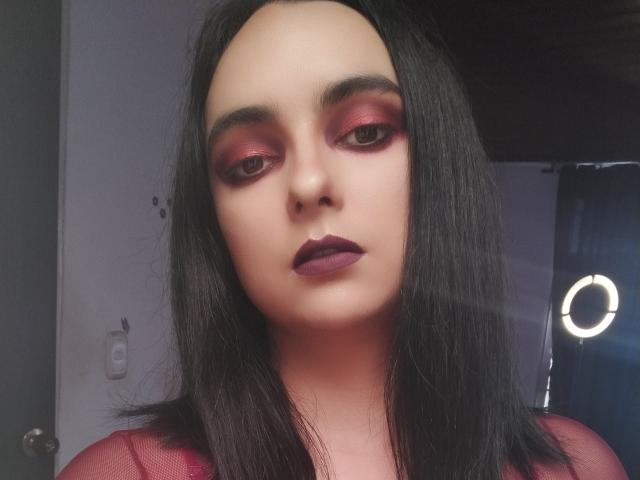 CandyParis has pale skin, dark black hair, and is wearing bold red eyeshadow, matching lipstick, and a sheer red top. Her tits are small, and she's looking straight at the camera.