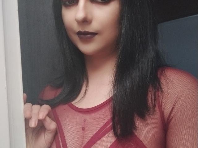 CandyParis com cabelo preto, batom escuro, vestindo uma blusa vermelha transparente, mostrando os seios, segurando o cabelo, em pé perto de uma porta..