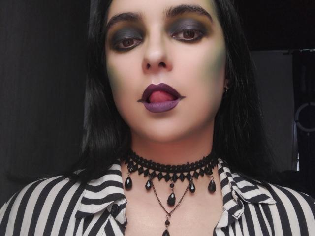 CandyParis has dark green and yellow eye makeup, purple lipstick, and black teardrop-shaped necklace. She's sticking out her tongue, wearing a striped black and white shirt.