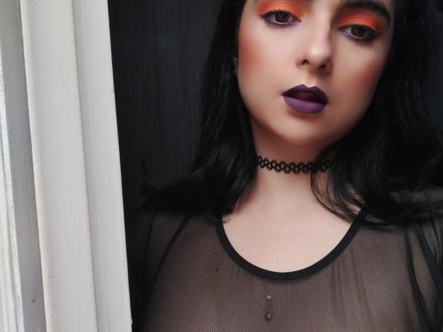 CandyParis met zwart haar, oranje oogschaduw, paars lippenstift, een doorschijnend zwart topje, een choker en zichtbare tepels, staand bij een wit gordijn..