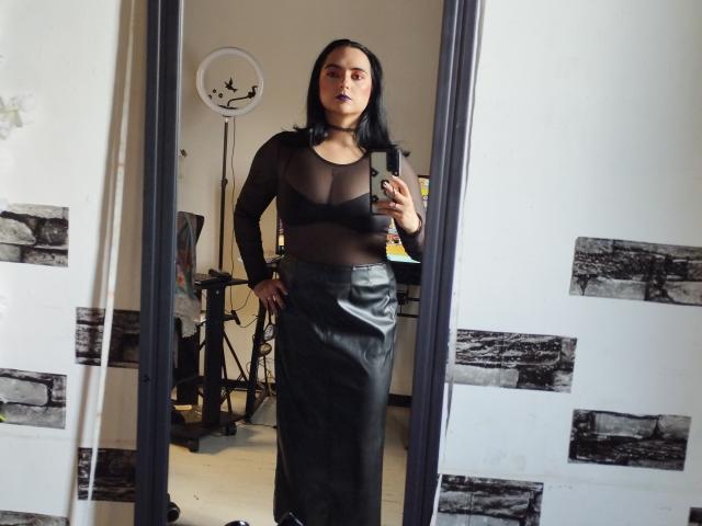 CandyParis with black sheer top, showing her small tits, and black leather skirt, taking a mirror selfie, holding a phone in a room with a ring light.