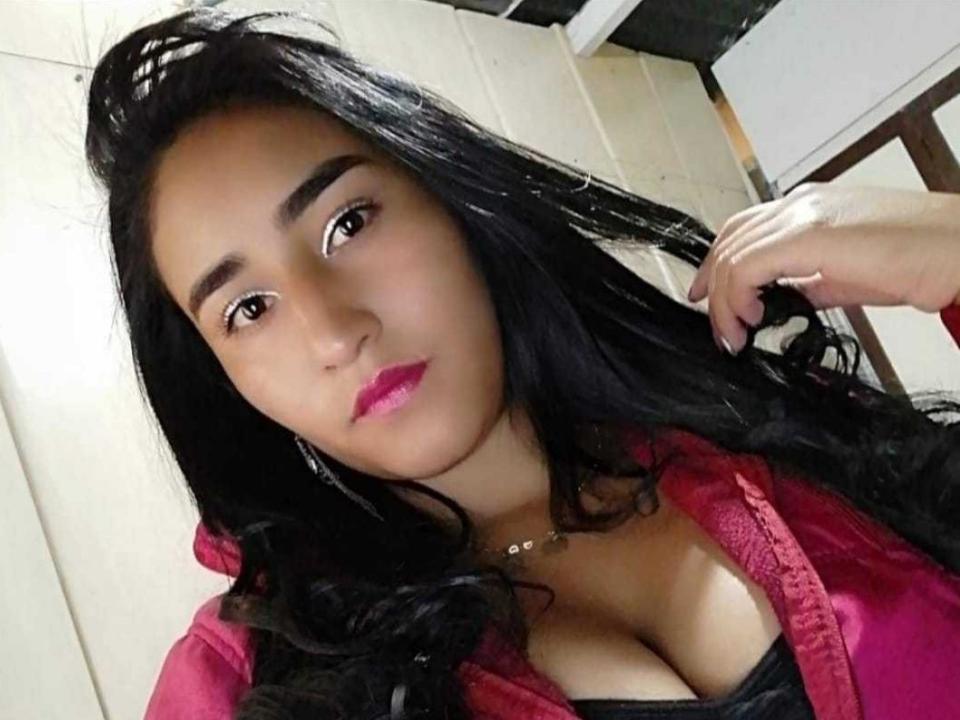 AmyLatiin with long black hair, pink lipstick, and a red jacket, holding her hair, showing cleavage in a black top, wearing a necklace, looking at the camera.