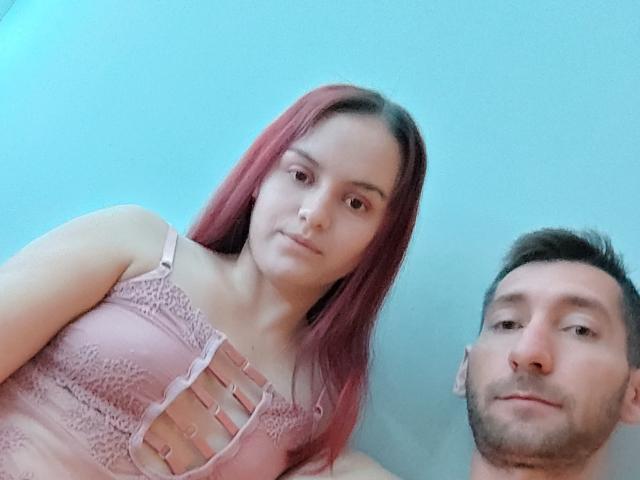 TeenyCouple with pink hair and lace pink top, next to a bearded man with short brown hair, both smiling. Her tits are covered by the lace top.