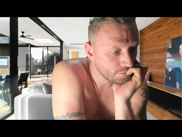 AlexMan69 is a shirtless, tattooed dude with a beard, thinking, resting his chin on his hand, and has a tattoo on his right arm. He looks slightly annoyed.