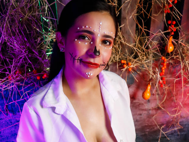 LailaRogerts with dark hair, white shirt, face painted with white dots, black nose, and cross lines, red lipstick, standing against colorful, tangled lights, and glowing ornaments.