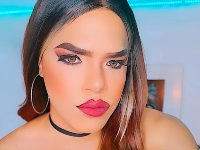 MinaYCelesteMistress has red lipstick, dark eyebrows, long eyelashes, hoop earrings, and a black choker, with brown hair and tan skin. Her brown eyes are looking forward.