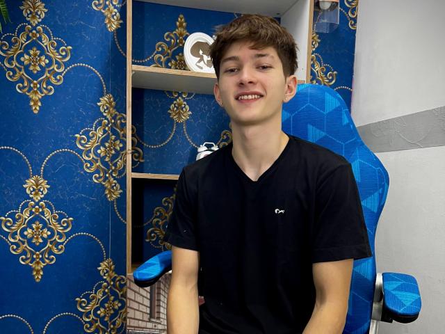 JakeDavins, young dude with short brown hair, black tee, and a smile, sitting in a blue hex-patterned chair, has a slim build and a cute face.