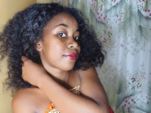 BleuSweety with curly black hair, brown skin, red lipstick, and a leopard print top, arms crossed, smiling slightly, looking at camera.