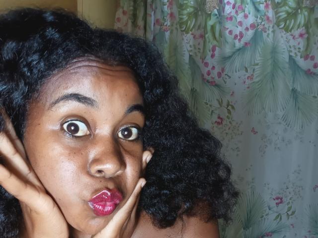 BleuSweety with curly black hair, wide eyes, and glossy pink lips, holding her face with hands, looking surprised. She's got a smooth brown skin tone and a cute face.