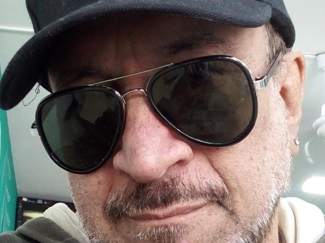 AmantsChauds ha un viso segnato, con una barba grigia, un berretto nero e occhiali da sole neri, e mostra un leggero sorriso. Il suo naso è prominente e porta un piccolo orecchino..