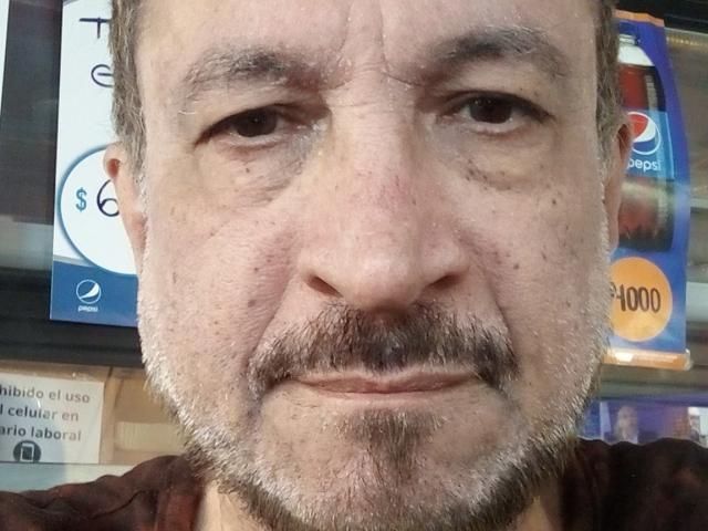 AmantsChauds ha una barba e dei baffi trasandati, una pelle leggermente segnata da rughe e un'espressione seria. Indossa una camicia marrone con un primo piano del suo viso..