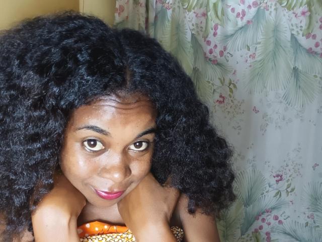 BleuSweety with curly black hair, brown eyes, and pink lipstick, leaning forward with hands near face, wearing an orange patterned top, showing her shiny dark brown tits.
