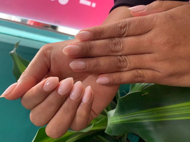 CarlaMuille's hands with pink, glossy, almond-shaped nails, resting on her dark brown skin, showing her long, manicured fingernails and gentle touch. Green leaf in background.