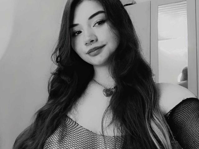 AmyWhitexd with long dark hair, nose ring, and fishnet top, showing her tits, wearing a heart necklace, smiling slightly, in a black-and-white photo.