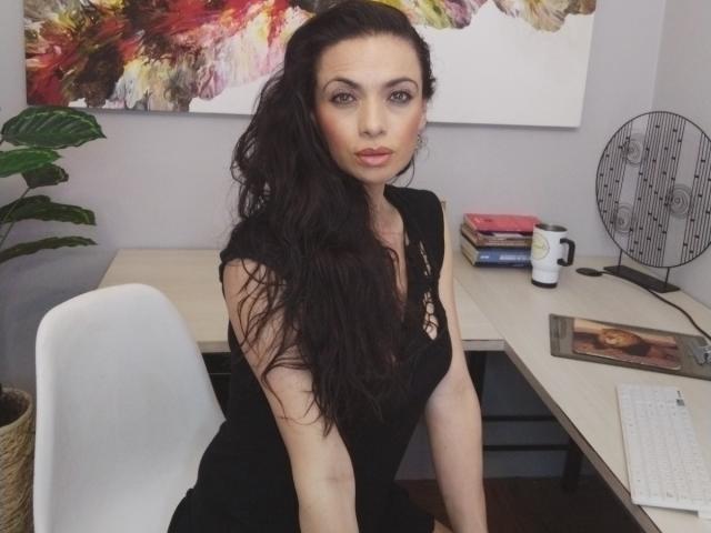 AfroditaRgenis, avec de longs cheveux noirs, portant une robe noire, assise à un bureau, regardant directement la caméra, les mains posées sur le bureau, chaise blanche, tasse de café et plante en arrière-plan..