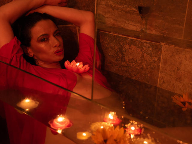 AfroditaRgenis in a red shirt, arms above head, in a candlelit bathtub with flowers, water, and a glass of wine. Her tits are exposed, looking seductive.