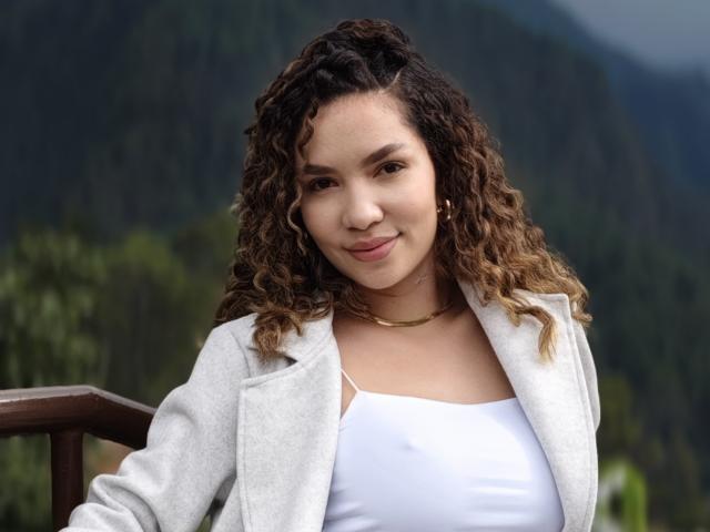 KendallPiierce69, con cabello castaño rizado, camiseta blanca y chaqueta gris, sonriendo a la cámara, llevando un collar dorado, de pie al aire libre, con montañas borrosas en el fondo..