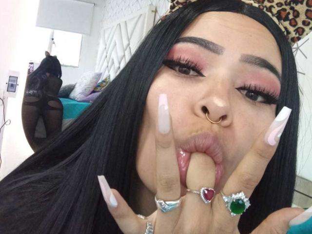MelodyBellanova with long black hair, pink eyeshadow, and long nails, sucking her fingers, wearing a nose ring and colorful rings, her mouth open, looking at the camera.