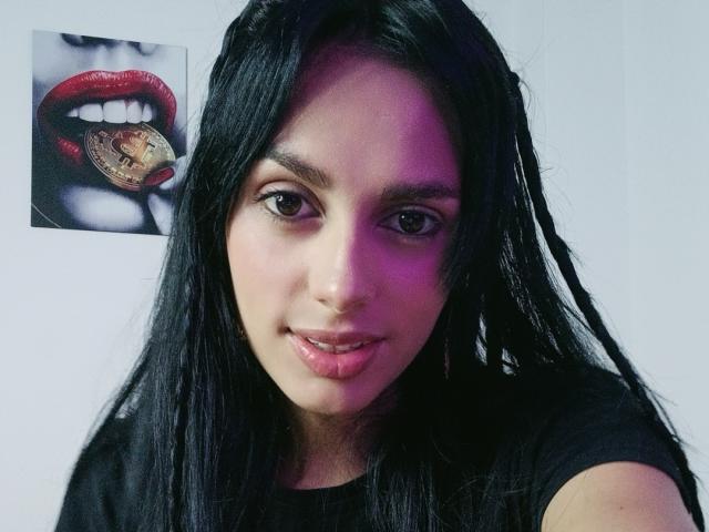 AnaisHilton with black hair, shiny pink lips, and brown eyes, wearing a black shirt, next to a photo of red lips with a gold coin in her mouth.