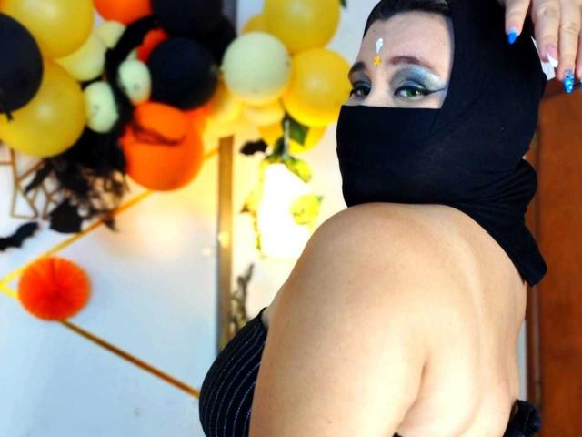 SalmaAzaman with blue eyeshadow, black mask covering her mouth and nose, black head covering, wearing a black strapless top, back to camera, yellow and black balloons in background.
