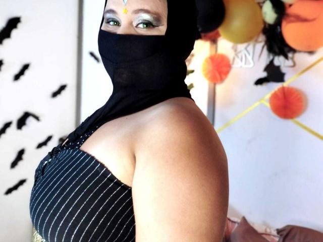 SalmaAzaman with dark pinstriped top, bare shoulders, black mask covering nose and mouth, green eyes, black head covering, visible tits, orange and black Halloween decorations in background.