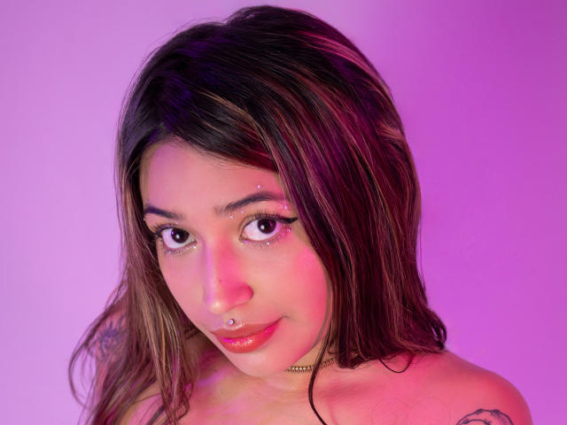 KatianPierce tiene el cabello castaño con reflejos rosados, un piercing en la nariz y lápiz labial rojo. Está mirando a la cámara con una pequeña sonrisa, mostrando sus tatuajes..