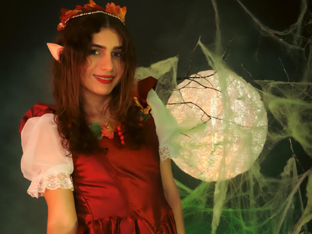 AndyDupont con orecchie a punta, vestito rosso, maniche di pizzo bianco, collana verde e fascia per capelli con un fiore rosso, sorride a una luna luminosa, ricoperta di ragnatele. Capelli castani di media lunghezza, pelle chiara..