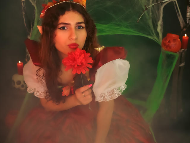 AndyDupont with curly brown hair, elf ears, red dress, white puffed sleeves, holding red flower to nose, sucking, green mist, red candles, skull, orange pumpkin, warm lighting.