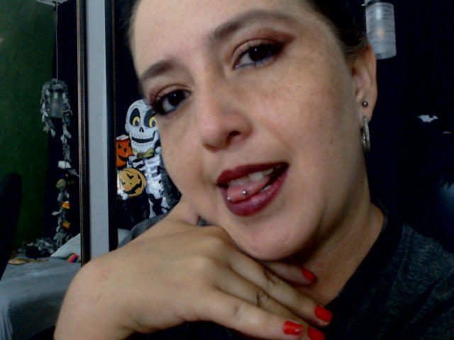 MaraLeon has freckled skin, dark hair in a bun, red lipstick, pierced tongue, red nail polish, and earrings, smiling with hand on chin in a selfie.