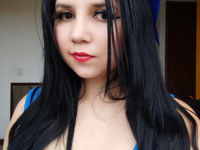 Kkatalina has long black hair, red lipstick, and blue top, with her tits slightly visible, looking at the camera with a slight smile, wearing a blue bow on her head.