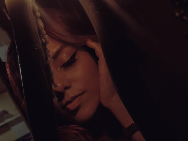 ZoeeMar, con los ojos cerrados, sombras oscuras, luz suave en su rostro, mano tocando la mejilla, maquillaje sutil, cabello castaño oscuro, nariz pequeña, expresión serena, tonos marrones cálidos, ambiente íntimo..