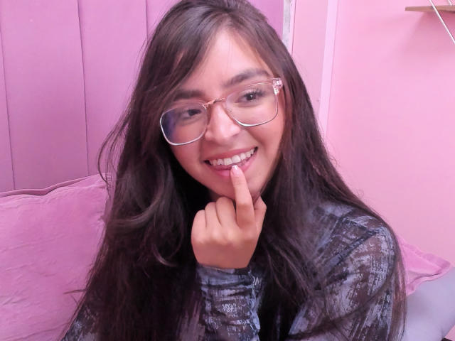 EvaMcAllister with long black hair, glasses, smiling, finger to lips, wearing a gray patterned shirt, pink room, pink wall, pink pillow, cute, sexy, playful, subtle.