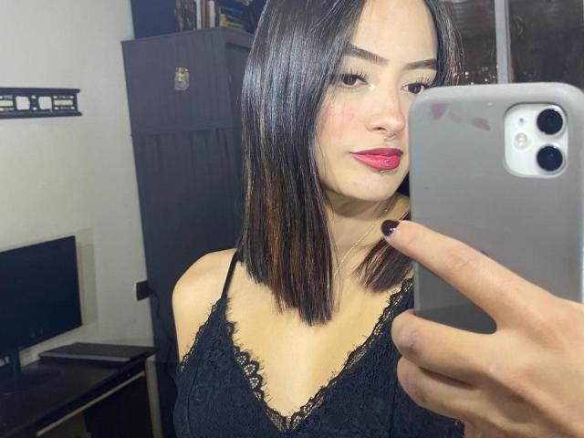 MiraCataleyax with shoulder-length black hair, red lipstick, black lace top, holding a phone, pointing at it, smiling, nose ring, black nails, in a room.