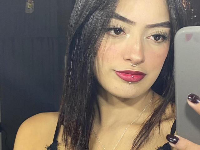 MiraCataleyax has dark brown hair, red lipstick, and a nose ring, holding a phone with black nails, looking into the mirror with a slight smile.