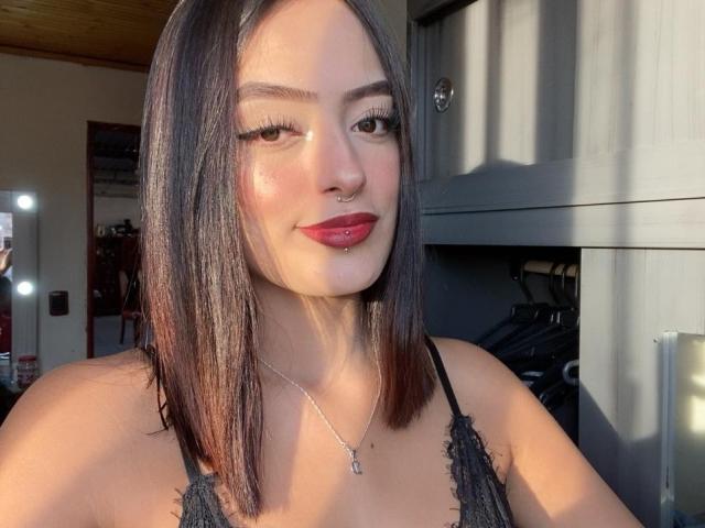 MiraCataleyax with medium-length brown hair, red lipstick, black lace top, nose ring, and long eyelashes, smiling at the camera in bright sunlight. Boobs visible, necklace with small cross.