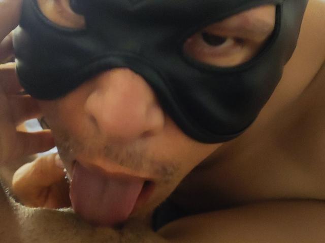 ApoloniaAndRocco met een zwart masker, tong uit, die een behaarde penis likt. Het gezicht van ApoloniaAndRocco is dichtbij, de ogen half open, en toont een enigszins vuile, intieme handeling..