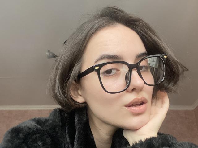 EstellaElsie with short brown hair, black glasses, and a black fuzzy jacket, resting her chin on her hand, looking slightly to the side with parted lips.